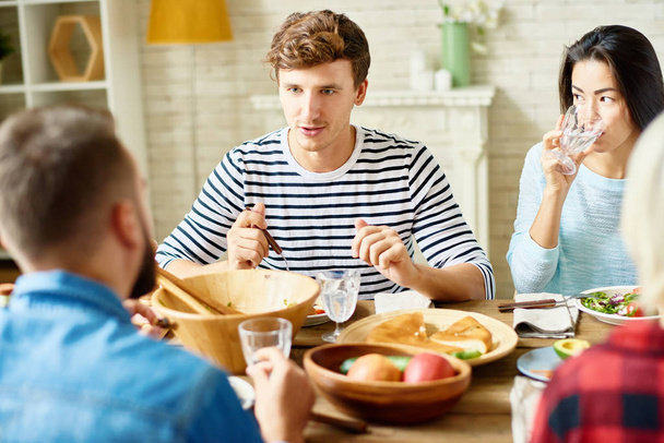 Portrét moderní mladý muž těší večeře s přáteli, posezení u velkého stolu s jídlem a mluví s lidmi - Fotografie, Obrázek