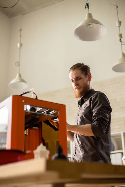 Low angle view of handsome bearded designer using 3D printer while working on ambitious project, interior of modern studio on background - Fotoğraf, Görsel