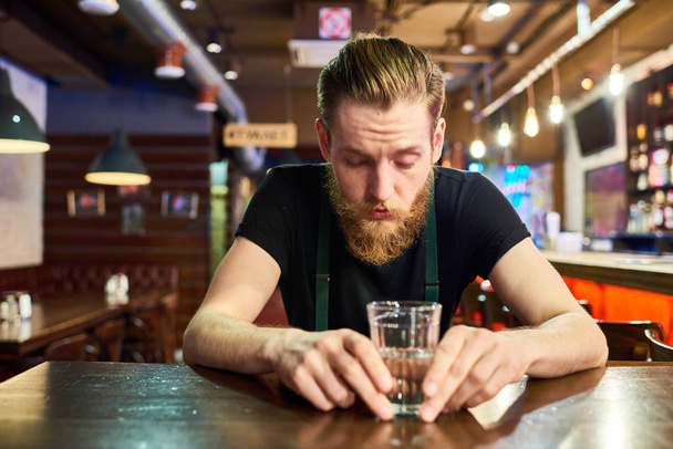 Retrato de vista frontal del hombre barbudo moderno emborrachándose solo en el pub sentado a la mesa y bebiendo vodka
 - Foto, Imagen