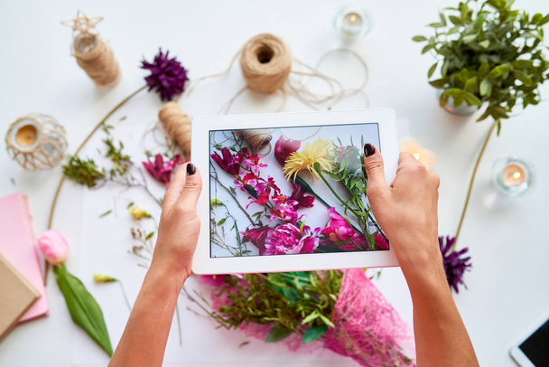 Top view closeup of female hands holding digital tablet and taking photo of floral decor composition for social media - Foto, afbeelding