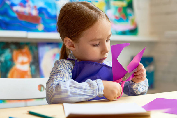 Portrait de mignonne petite fille découpant soigneusement les formes du cœur tout en faisant une carte faite à la main pendant les cours d'art et d'artisanat à l'école maternelle
 - Photo, image