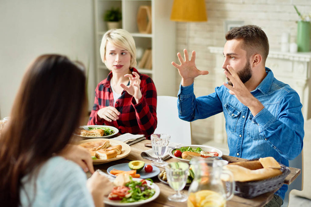 Portrait of young friends enjoying dinner together and big festive table and telling stories gesturing actively in lively conversation - Photo, image