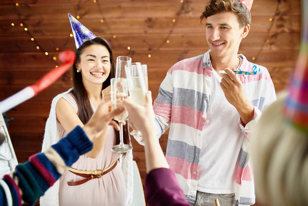 Grupo de jóvenes felices que usan gorras navideñas celebrando el cumpleaños con amigos sacudiendo copas de champán durante la fiesta en casa, se centran en la pareja sonriente
 - Foto, Imagen