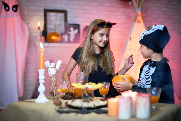 Muotokuva kaksi lasta, poika ja tyttö yllään Halloween puvut chattailuun aikana puolue seisoo pöydässä makeisia ja välipaloja koristeltu huone
 - Valokuva, kuva