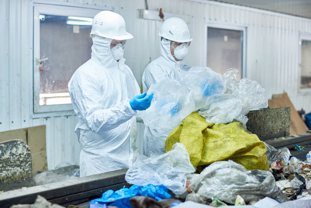 Portrait de deux travailleurs portant des combinaisons à risque biologique dans une usine de traitement des déchets triant le plastique recyclable et le carton sur une bande transporteuse
 - Photo, image