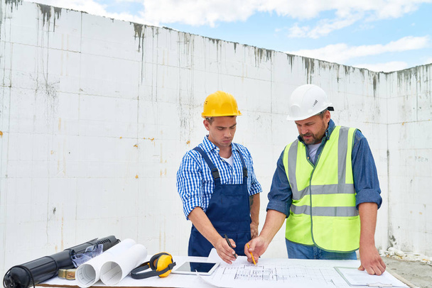 Portrait de deux travailleurs de la construction discutant des plans d'étage et de la documentation technique sur place, faisant des marques sur les plans directeurs
 - Photo, image