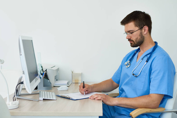 Side view portrait of experienced doctor  sitting at desk in doctors office filling in patient forms  against white background - Photo, Image