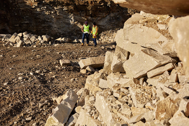 Amplio ángulo de tiro de dos trabajadores industriales con chaquetas reflectantes, uno de ellos africano, caminando en el sitio de trabajo minero al aire libre, se centran en la pila de rocas en primer plano, espacio de copia
 - Foto, imagen