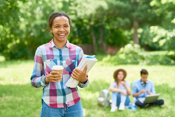 Retrato de un estudiante feliz de raza mixta parado al aire libre en el césped verde sosteniendo libros y sonriendo mirando a la cámara con otras personas en el fondo
 - Foto, Imagen