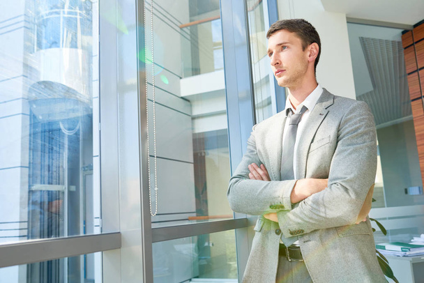 Portrait of confident young businessman standing by window with arms crossed looking away pensively, copy space - Foto, afbeelding