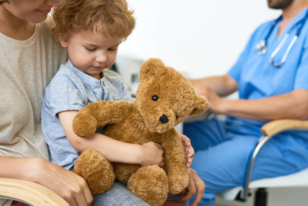 Πορτρέτο του χαριτωμένο μικρό παιδί που κάθεται στην αγκαλιά μητέρες στο γραφείο ιατρών σε αναμονή για έλεγχο αγκάλιασμα παιχνιδιών βελούδου αρκουδάκι - Φωτογραφία, εικόνα