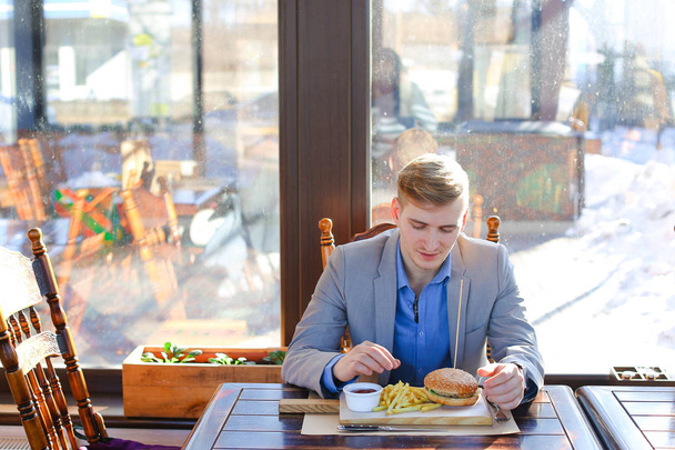 Retrato del joven hombre de negocios ocupado sentado con un vaso de bebida y hamburguesa en el interior del restaurante de comida rápida. Atractivo hombre caucásico comiendo y utilizando la computadora portátil en la cafetería. En interiores
 - Foto, imagen