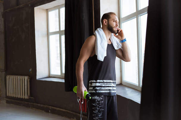 Deportivo hombre activo que se queda cerca de la ventana en el gimnasio con el móvil en la mano y mirando lejos. Charlando por teléfono
. - Foto, imagen