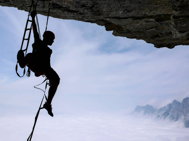 male rock climber aid climbing a difficult technical overhanging roof with a fantastic view of mountains behind and a sea of clouds below - Photo, Image