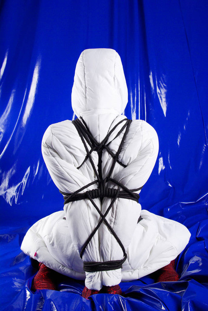 strange and cute sex game with bondage by black ropes of slim woman in a slinky fetish catsuit and white down jacket - Photo, Image