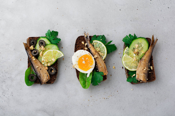 Fish sandwiches with sprats, cucumber, lime, boiled eggs, parsley leaves and mango on rye bread on a gray old concrete or stone background. Selective focus. Rustic style. Top view. Copy space. - Zdjęcie, obraz