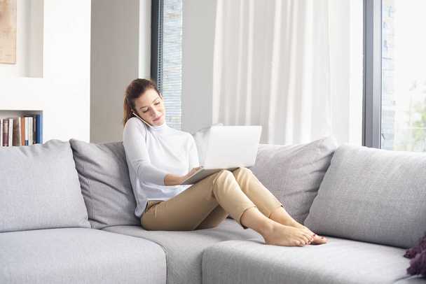 Full length of attractive middle aged woman using her cell phone and making call while sitting on sofa with laptop and working from home office. - Photo, Image