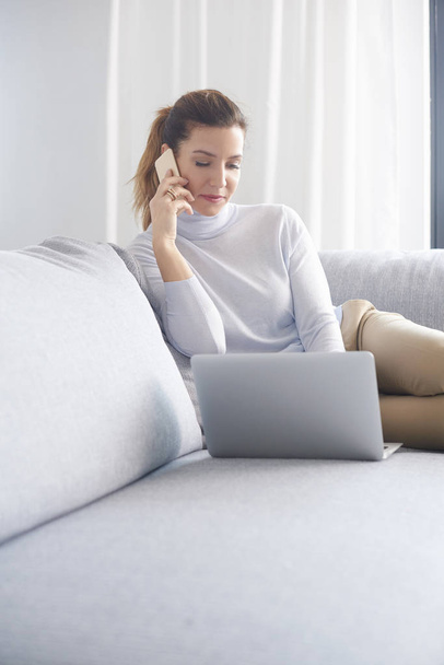 Smiling attractive woman using mobile phone and making call while sitting on sofa with laptop and working from home. Home office.  - Fotoğraf, Görsel