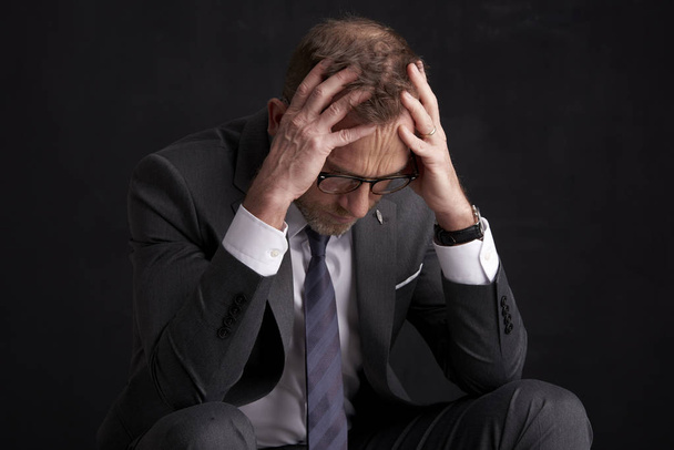 Stressful middle aged businessman in suit sitting at dark background with hands on his forehead. - Foto, afbeelding