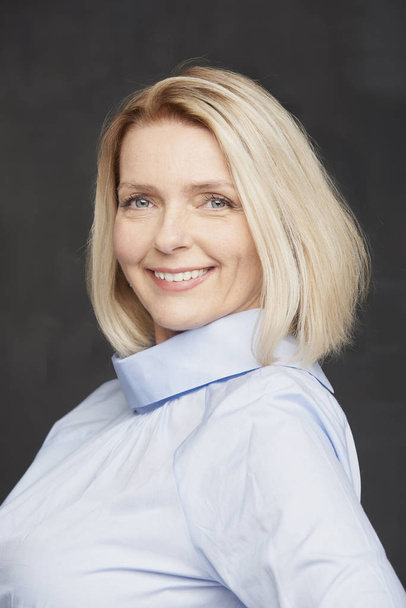Beautiful blonde smiling mature woman in light blue blouse posing before dark wall - Foto, Imagen