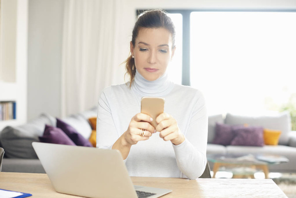 Привлекательная женщина с помощью мобильного телефона и текстовых сообщений во время работы на ноутбуке из дома
. - Фото, изображение