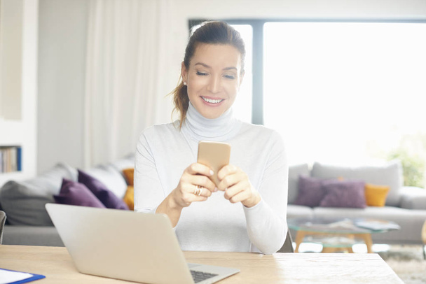 Happy woman using her mobile phone and text messaging while working on laptop from home. - Фото, изображение