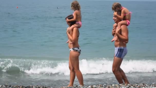 man and woman holding little girls on shoulders walks on beach with sea surf - Záběry, video
