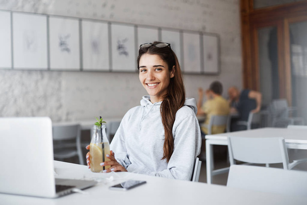 Cute attractive european young woman sitting in front of laptop, wearing trendy sweatshirt and stylish glasses, drinking beverage while in cafe. Freelancer took break from work, sipping cocktail - Foto, Bild