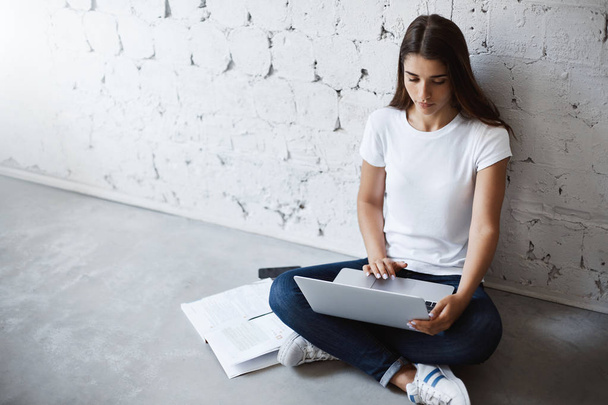 Young attractive trendy student sitting on floor with crossed feet, leaning on wall and working in laptop, surrounded with notes. Young teacher prepares for next lesson in language school - Photo, image