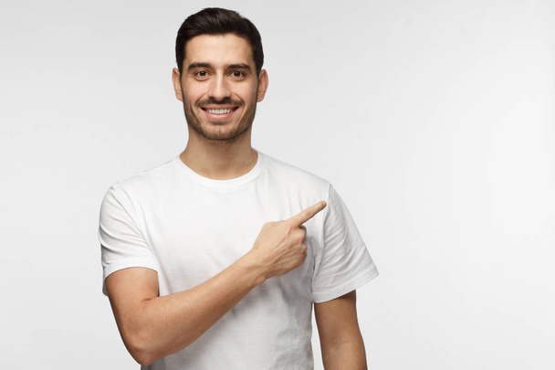 Attractive young man in white t-shirt pointing right with his finger isolated on gray background - Фото, изображение