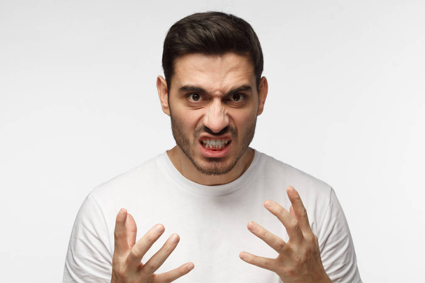 Shut up concept. Mad young man bite his teeth, gesturing with hands. Studio portrait of angry and irritated male in white tshirt yelling, looking crazy and full of anger - Фото, изображение