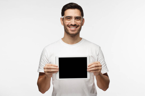 Studio picture of positive man isolated on grey background standing in casual clothes holding tablet and showing it blank screen with happy smile as if advising product or service - Zdjęcie, obraz
