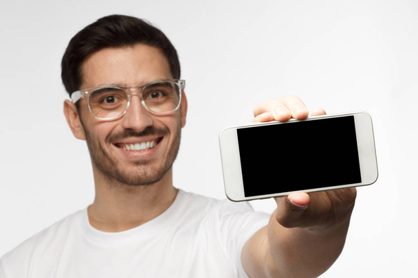Closeup of young handsome man smiling isolated on grey background, holding and showing horizontal blank smartphone with copyspace for advertisement of applications, websites, goods and services - Fotoğraf, Görsel