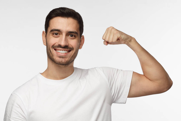 Cheerful smiling athletic man in white tshirt flexing bicep and looking at camera isolated on the gray background - Фото, зображення