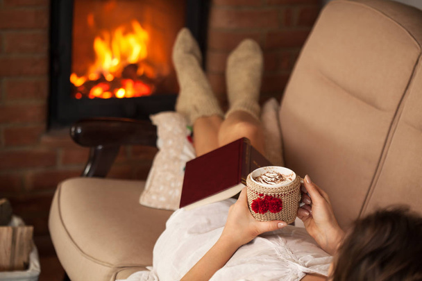 Mulher relaxante pelo fogo segurando uma xícara de chocolate quente com c
 - Foto, Imagem