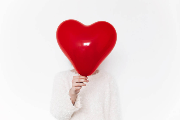 woman in white sweater holding red balloon heart hiding her head standing on white background - Valokuva, kuva