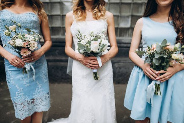 Γάμου. Η νύφη σε ένα λευκό φόρεμα στέκεται με παράνυμφοι της που βρίσκονται σε παστέλ μπλε φορέματα, όλοι κατέχουν ανθοδέσμες στα χέρια τους - Φωτογραφία, εικόνα