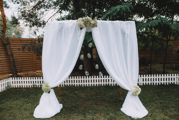 Свадебная арка с белой тканью и цветами в минималистском стиле в саду
 - Фото, изображение