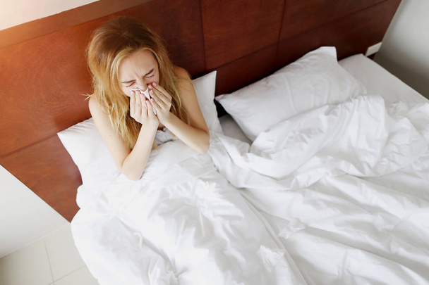 Κορίτσι είναι άρρωστο, φτερνίζεται στο λευκό κρεβάτι με χαρτοπετσέτα στο σπίτι ηλιόλουστη μέρα - Φωτογραφία, εικόνα
