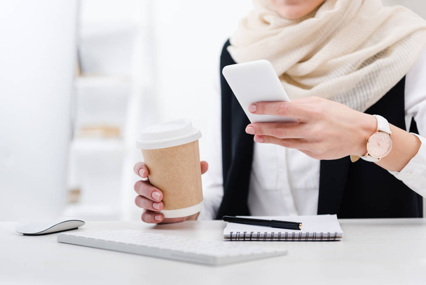 partial view of businesswoman with coffee to go using smartphone at workplace - Φωτογραφία, εικόνα