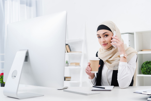 businesswoman in hijab with coffee to go talking on smartphone at workplace in office - Foto, immagini