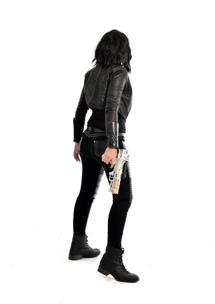 po celé délce portrét černé vlasy dívka, která nosí kožené oblečení. stojící pozice zatímco drží zbraň, izolované na bílém studio pozadí. - Fotografie, Obrázek