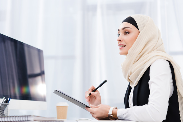 side view of smiling businesswoman in hijab with documents at workplace in office - Фото, изображение