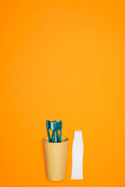 top view of toothbrushes in paper cup and tube of toothpaste, isolated on orange - Fotó, kép