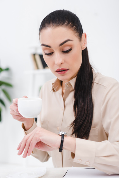 portrait of businesswoman with cup of coffee checking time at workplace in office - Foto, Imagem