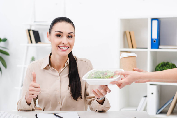 Portret van Glimlachende zakenvrouw weergegeven: duim omhoog houdend voedsel uit courier in office meenemen - Foto, afbeelding