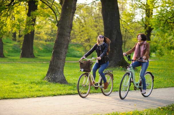 Two young attractive women ride bikes in the spring park. Pleasure by nature and movements. Healthy lifestyle. - 写真・画像