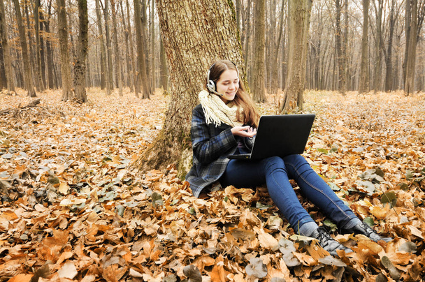 Beautiful young girl with headset working on black laptop in park with fallen leaves during autumn season - Valokuva, kuva