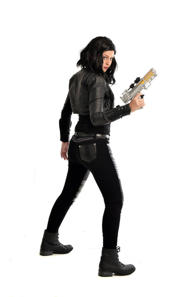 po celé délce portrét černé vlasy dívka, která nosí kožené oblečení. stojící pozice zatímco drží zbraň, izolované na bílém studio pozadí. - Fotografie, Obrázek