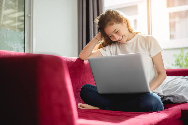 Νεαρή γυναίκα ψώνια σε απευθείας σύνδεση με το internet για χαρούμενη διάθεση σε κόκκινο καναπέ. Επιχειρήσεις και εργάζονται έννοια, χαλαρώστε και μέρος του χρόνου της έννοιας - Φωτογραφία, εικόνα